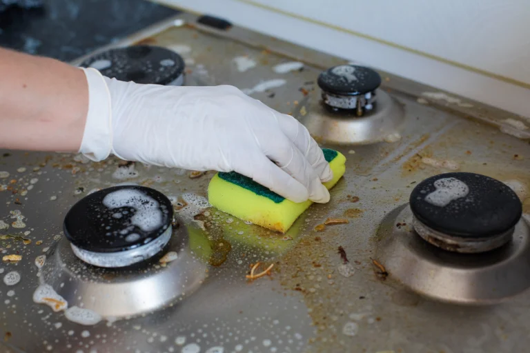 Mão feminina usando uma luva de látex enquanto limpa um fogão engordurado com uma esponja, ilustrando o artigo da SuperPro Bettanin sobre desengordurante profissional.