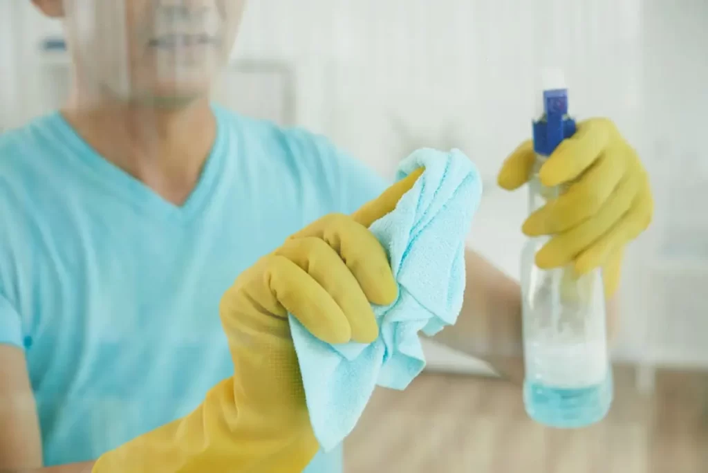 Homem com luvas de limpeza segurando pano microfibra e borrifando produto em vidro