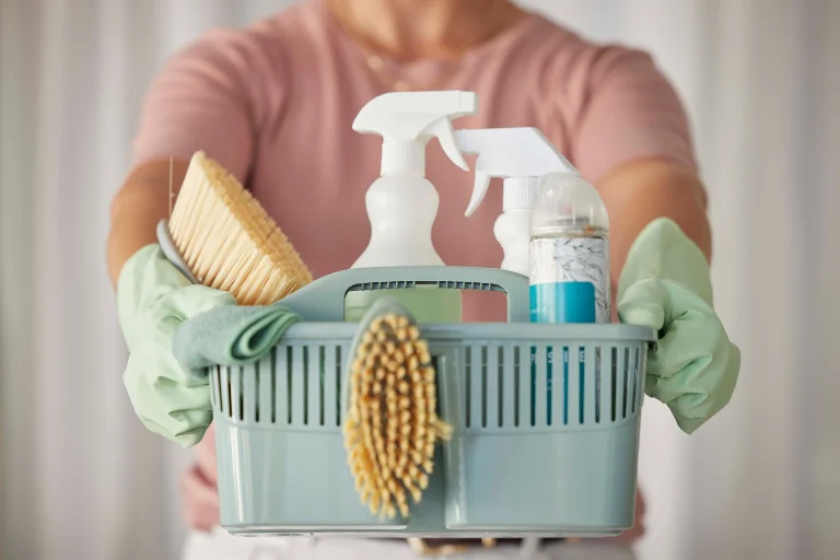 Mãos femininas com luvas verdes seguram um cesto com produtos de limpeza, ilustrando um artigo da SuperPro Bettanin sobre lista de produtos de limpeza.