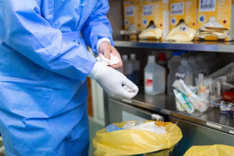 Profissional de saúde vestindo traje de proteção azul e colocando luvas brancas em ambiente hospitalar com equipamentos médicos ao fundo, ilustrando um artigo da SuperPro Bettanin sobre resíduos hospitalares.