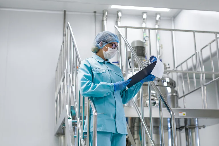 Profissional de laboratório com jaleco azul, touca, máscara e luvas segurando prancheta em ambiente industrial de produção, representando um artigo da SuperPro Bettanin sobre ISO 9001.