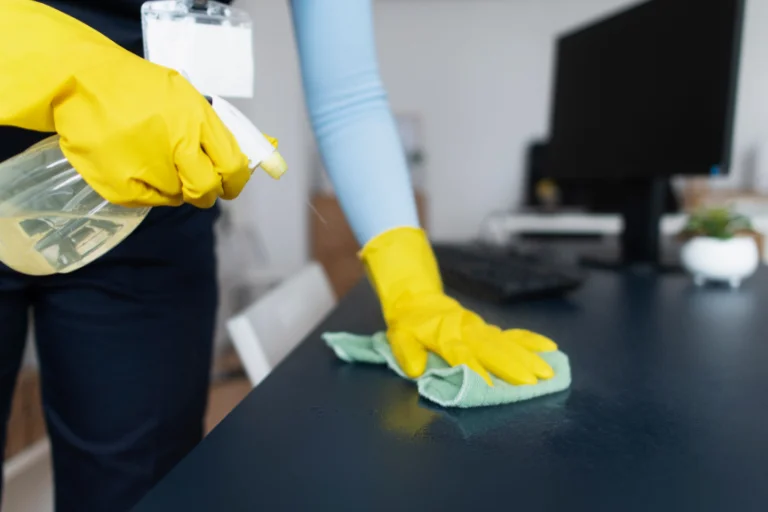 Pessoa limpando mesa de escritório com spray de limpeza e pano verde usando luvas amarelasPessoa limpando mesa de escritório com spray de limpeza e pano verde usando luvas amarelas, ilustrando um artigo da SuperPro Bettanin sobre pilares fundamentais da higiene ambiental em empresas.