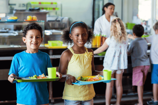 Duas crianças sorridentes carregando suas bandejas de comida em um refeitório escolar