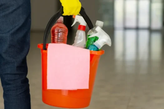Mão com luva amarela segurando balde laranja com produtos de limpeza e pano rosa, em artigo da SuperPro Bettanin sobre como comprar produtos de limpeza direto da fábrica.
