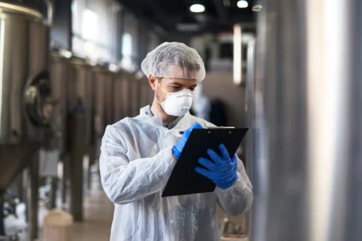 Homem com uniforme de segurança segura uma prancheta no meio de uma indústria, enquanto faz uma análise de higiene e segurança alimentar.