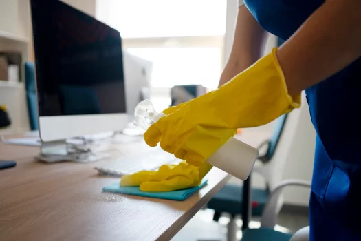 Pessoa limpando uma mesa de escritório com um pano e produtos de limpeza, usando luvas amarelas, ambiente bem iluminado.
