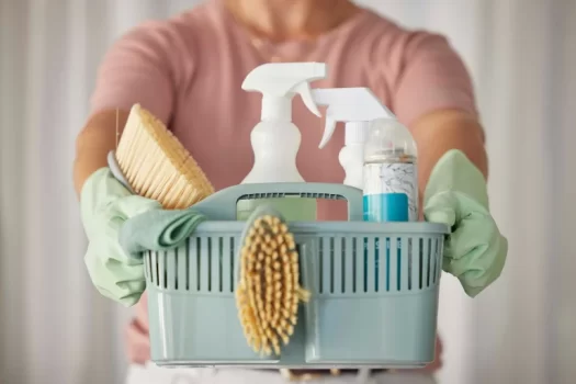 Mãos femininas com luvas verdes seguram um cesto com produtos de limpeza, ilustrando um artigo da SuperPro Bettanin sobre lista de produtos de limpeza.
