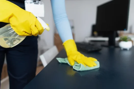 Pessoa limpando mesa de escritório com spray de limpeza e pano verde usando luvas amarelasPessoa limpando mesa de escritório com spray de limpeza e pano verde usando luvas amarelas, ilustrando um artigo da SuperPro Bettanin sobre pilares fundamentais da higiene ambiental em empresas.
